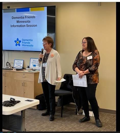 A photo of two people standing at the front of a room. Behind them is a projected screen with Dementia Friends information visible.